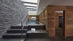 Un escalier intérieur moderne, doté d’une balustrade en verre, mène à un espace de détente avec un mur en bois et une vue sur l’extérieur.