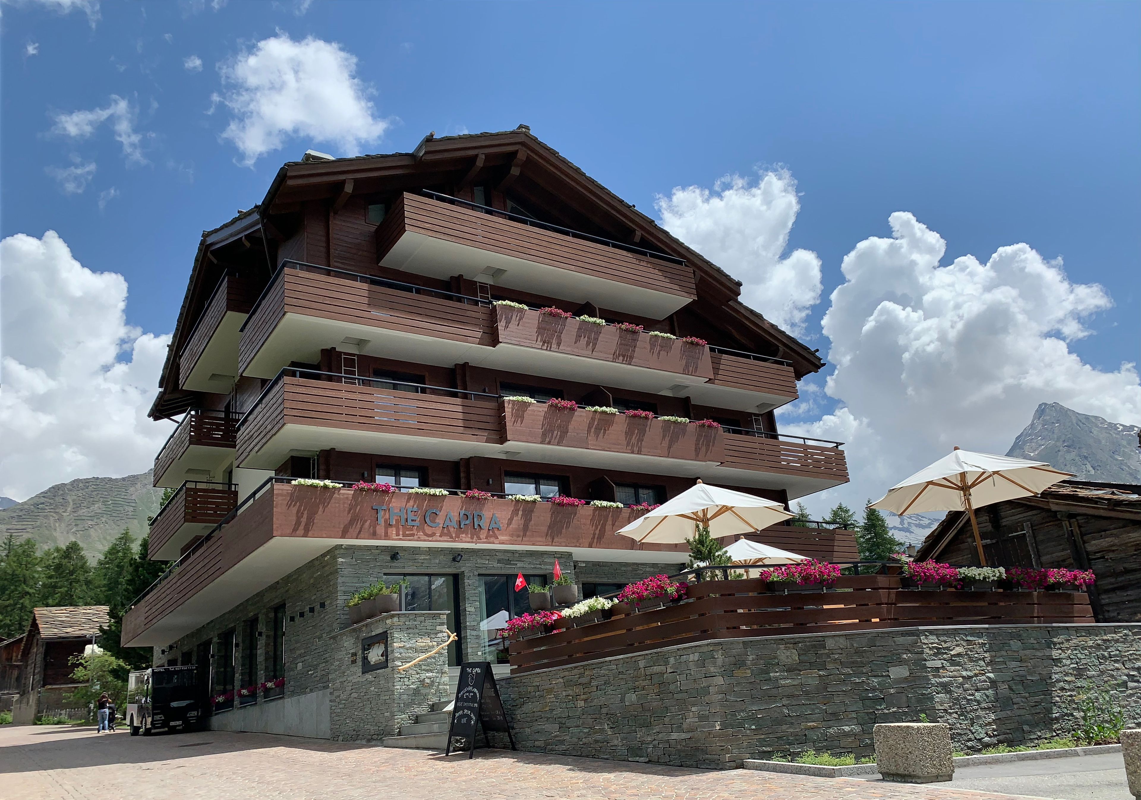 Un hotel de varios pisos con balcones decorados con flores, fachada de piedra y un letrero que dice “THE CAPRA”.