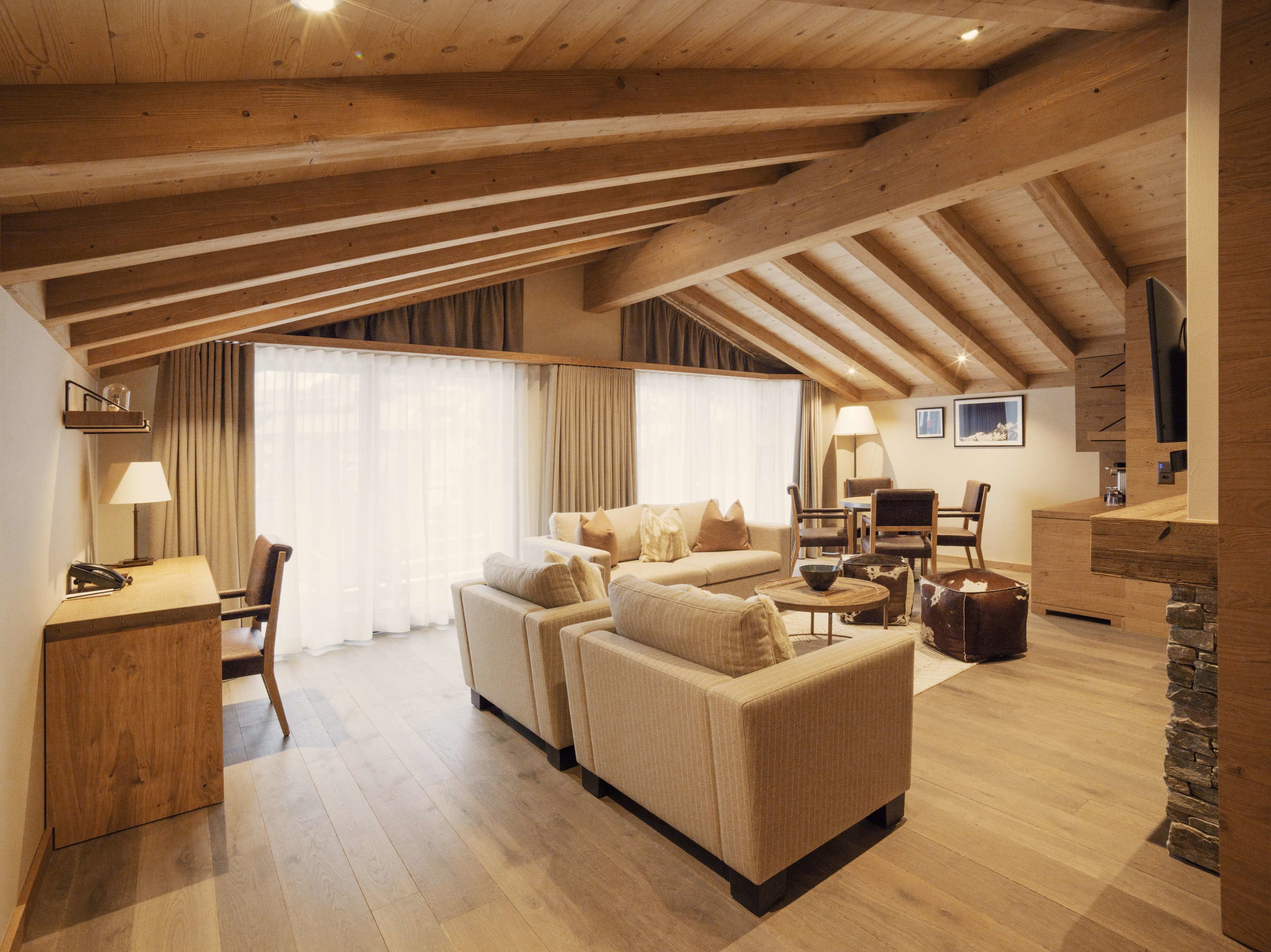 A spacious living area with a high ceiling, wooden beams, a sofa set, a dining table, and a TV mounted on the wall.