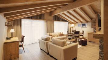 A spacious living area with a high ceiling, wooden beams, a sofa set, a dining table, and a TV mounted on the wall.