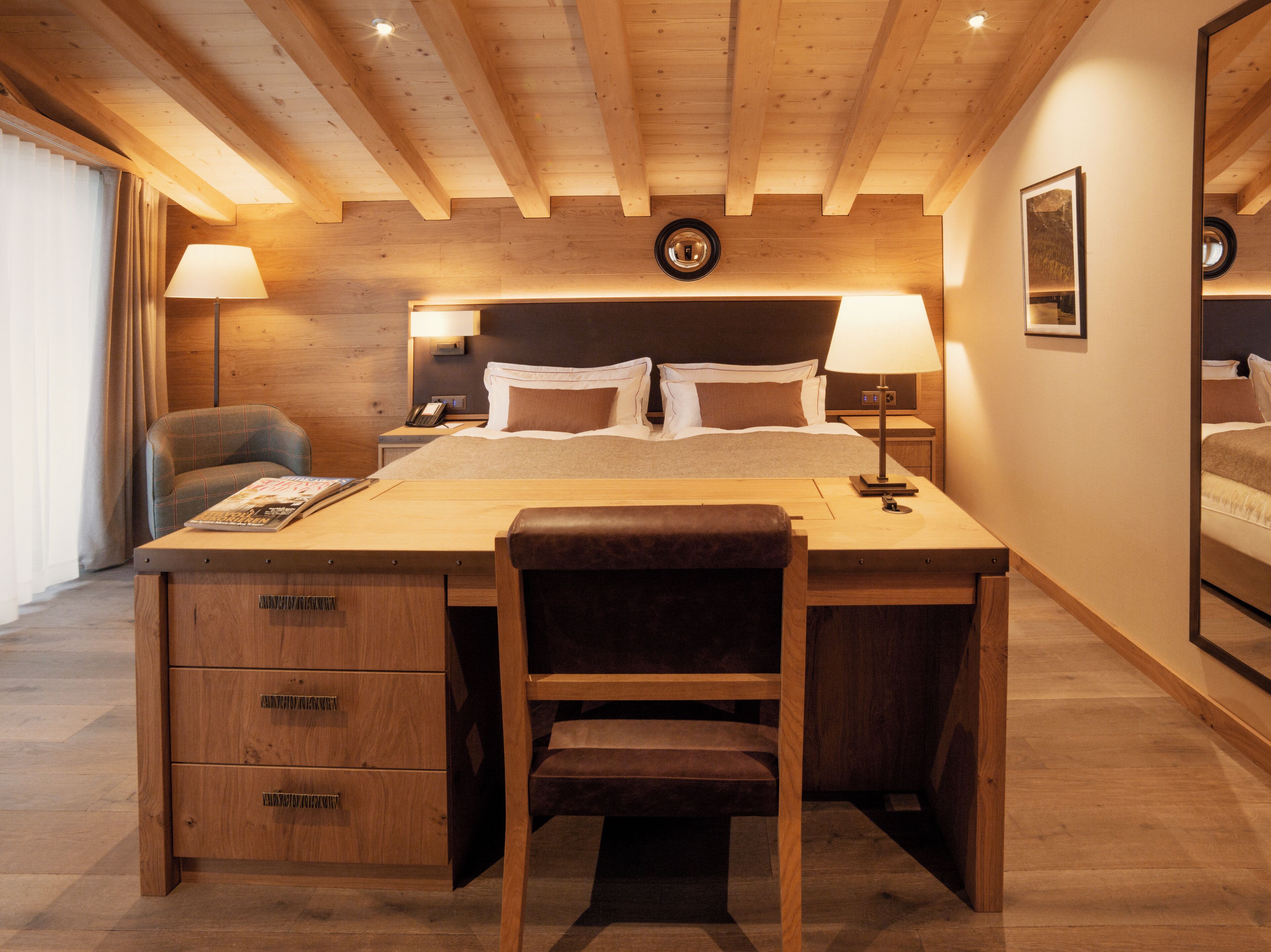 A hotel room with a wooden desk, a chair, a bed with pillows, a nightstand with a lamp, and a wall-mounted clock.