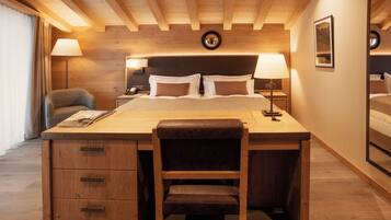 A hotel room with a wooden desk, a chair, a bed with pillows, a nightstand with a lamp, and a wall-mounted clock.