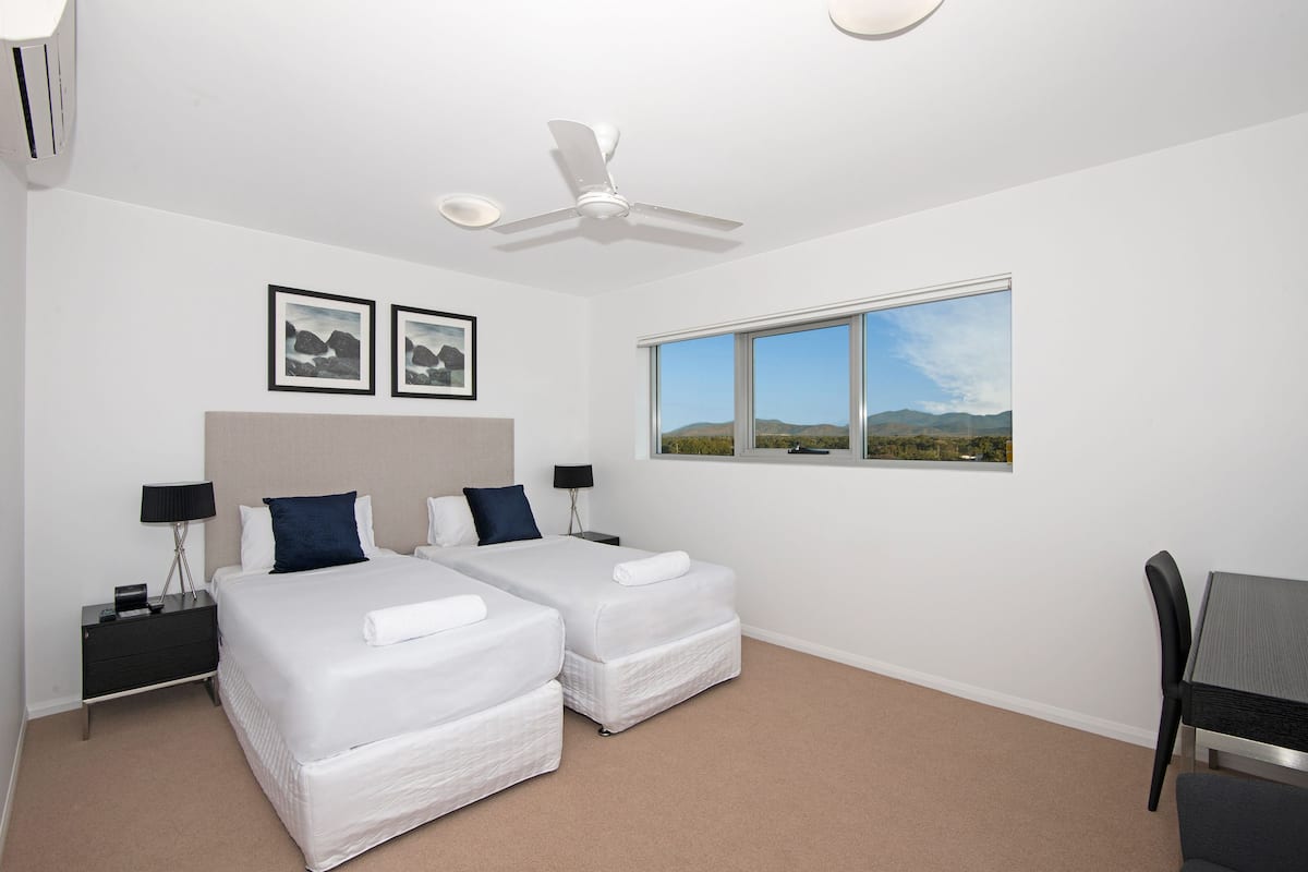 Habitación de hotel con dos camas, ventilador de techo, ventana con vista al paisaje y escritorio con silla.
