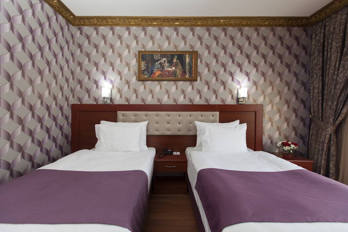 A hotel room with two beds, a wooden headboard, a painting on the wall, and a nightstand with a lamp and flowers.