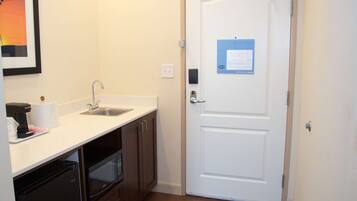 A hotel room with a kitchenette, including a sink, microwave, and refrigerator. There is a white door with an emergency exit sign.