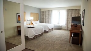 Habitación de hotel con dos camas, un escritorio, televisor y un ventanal grande con cortinas.