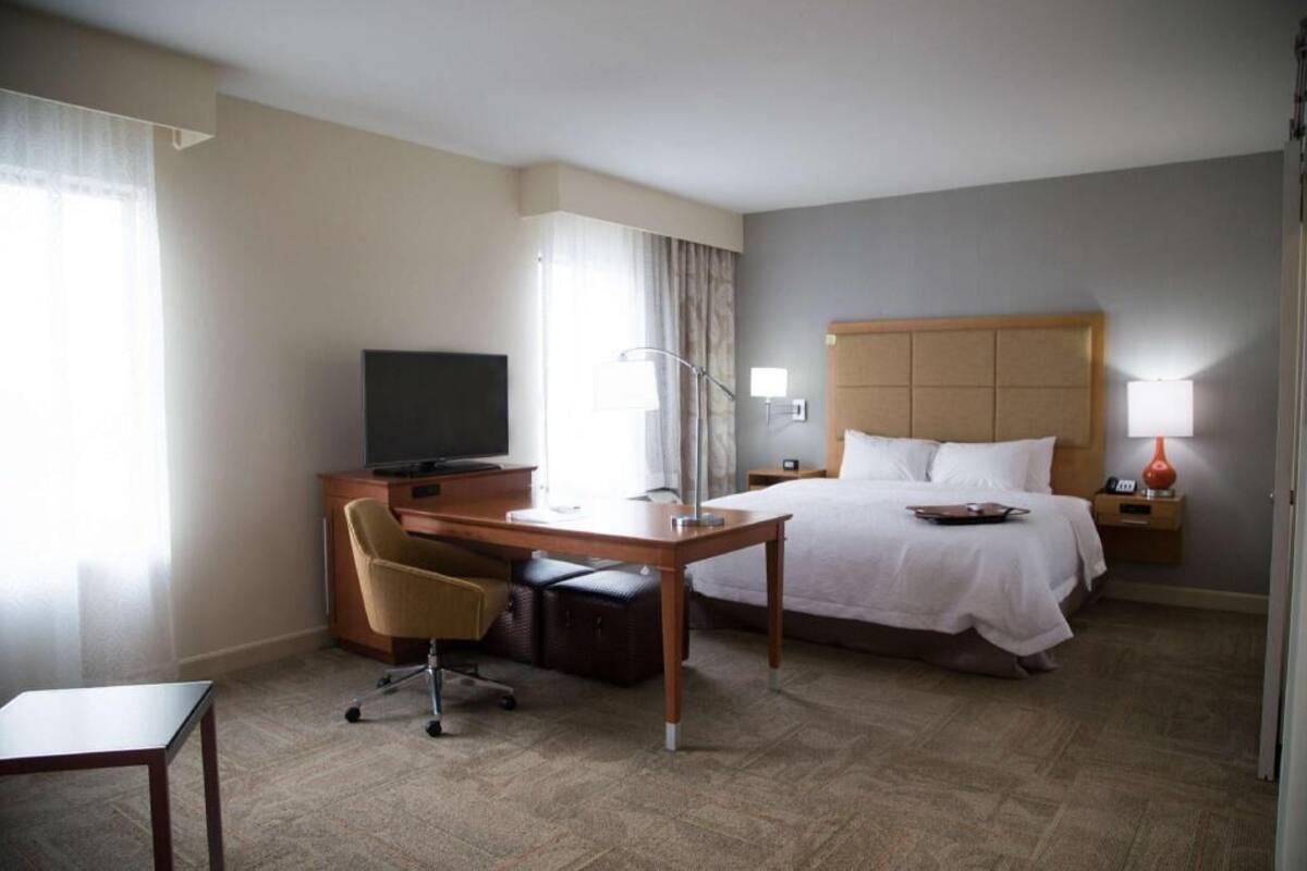 Quarto de hotel com cama, escrivaninha, cadeira, televisão, abajur e mesa de cabeceira.