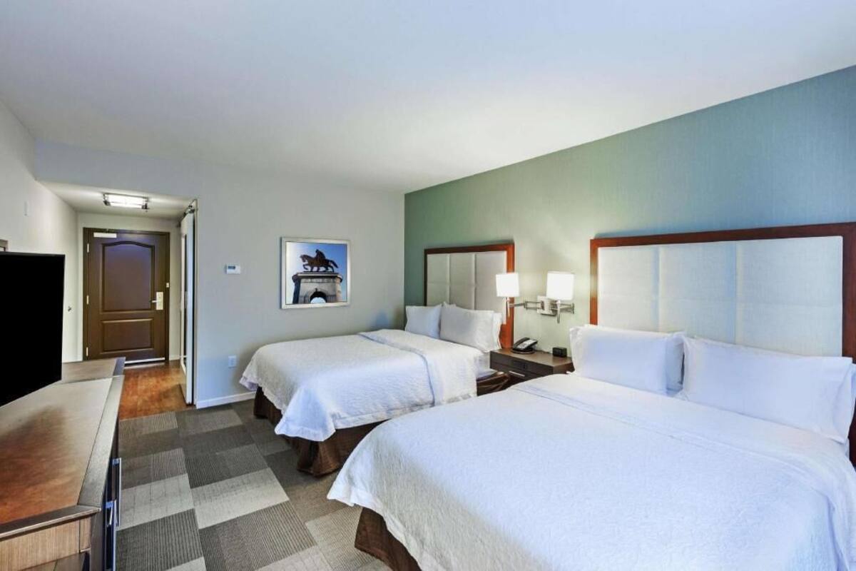 A hotel room with two beds, a desk, a chair, a lamp, and a poster on the wall.