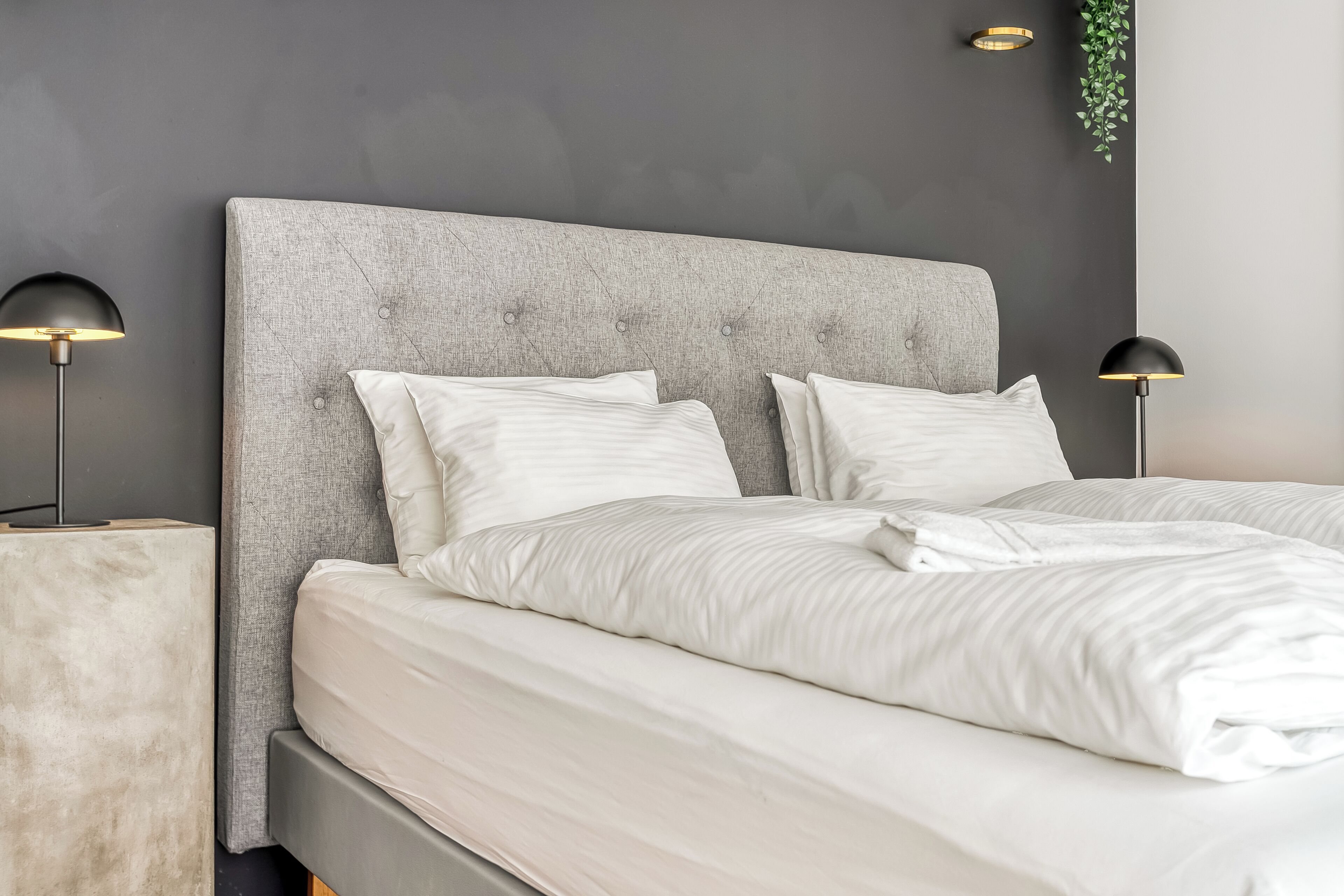 Un lit bien fait, doté d’une tête de lit rembourrée de couleur grise, de draps blancs et de deux lampes de chevet.