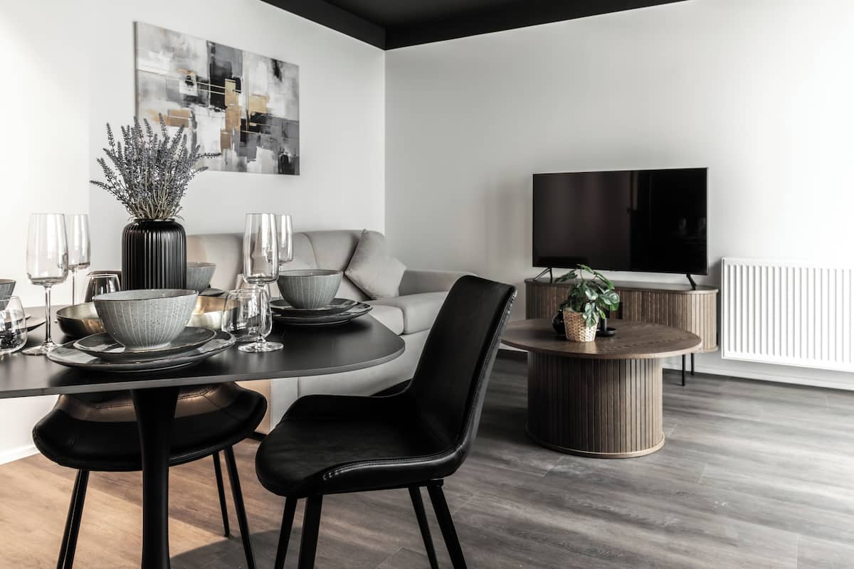 Un comedor moderno con una mesa redonda, sillas negras y un jarrón de flores. Un televisor de pantalla plana está montado en la pared, y se ve un sofá al fondo.