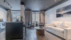 Un salon moderne avec un canapé, une table basse et des fauteuils. Il y a un coin cuisine équipé d’un réfrigérateur et d’un four à micro-ondes. La pièce est dotée de grandes fenêtres avec des rideaux.