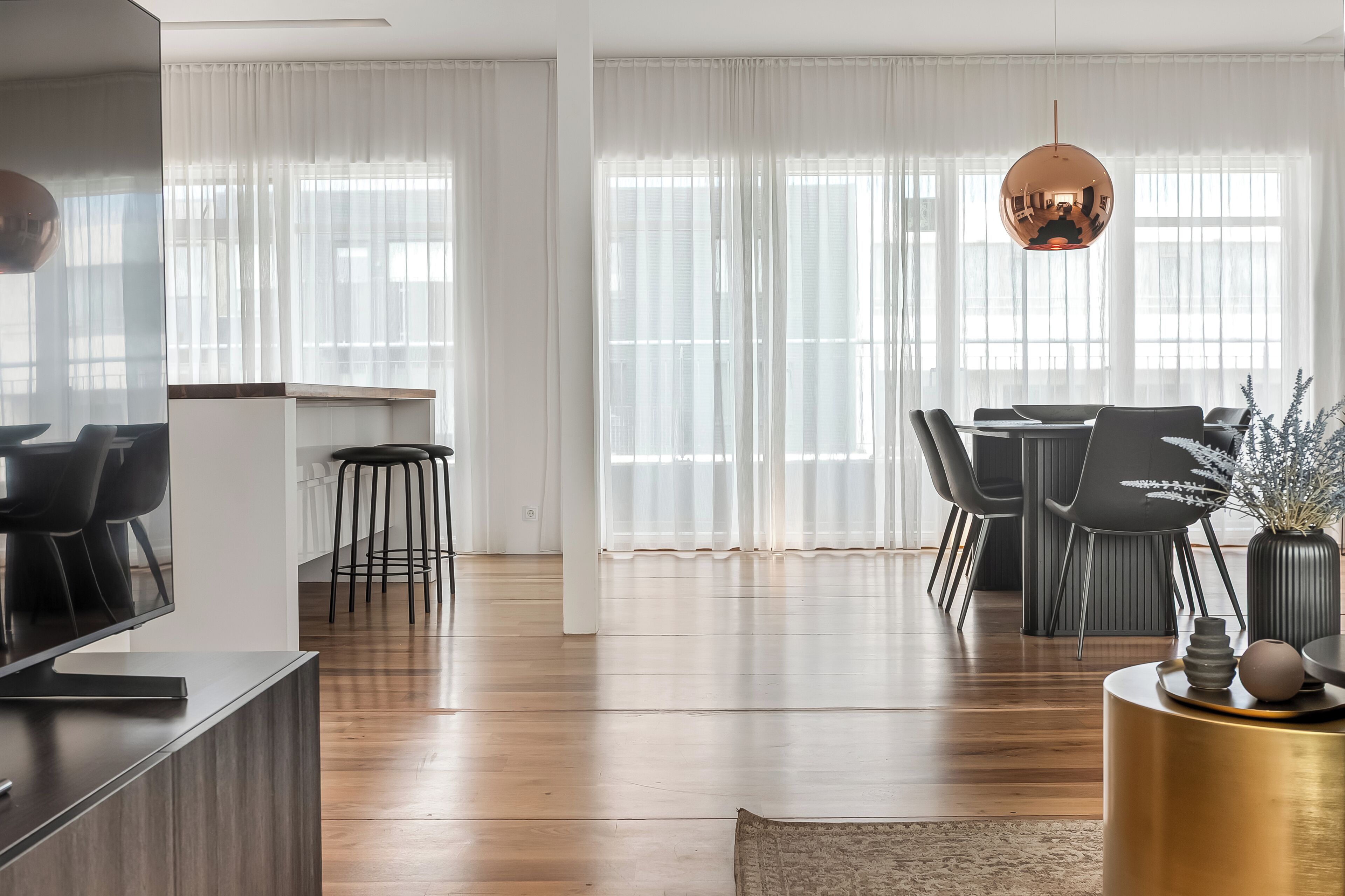 Una sala de estar moderna con comedor, suelo de madera, televisor y un ventanal con cortinas translúcidas.