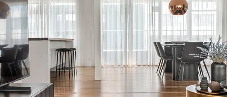 Una sala de estar moderna con comedor, suelo de madera, televisor y un ventanal con cortinas translúcidas.