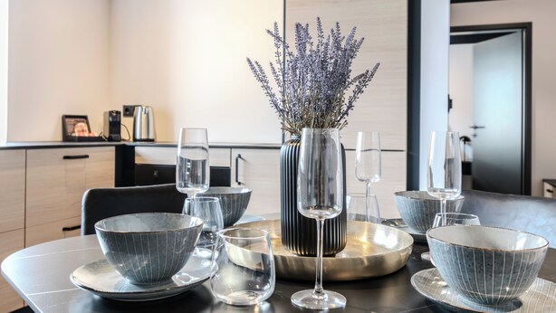 Un espace de salle à manger moderne, avec une table dressée pour quatre personnes, agrémentée de bols gris, de verres à vin et d’un vase contenant de la lavande.