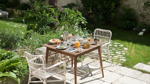 A wooden table set outdoors with white wicker chairs, surrounded by greenery and a stone wall. - Chambres d'Hôtes Eden Ouest (La Rochelle)
