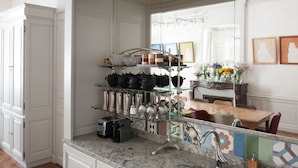 A kitchen with white cabinetry, a marble countertop, and a glass shelf displaying kitchenware. - Chambres d'Hôtes Eden Ouest (La Rochelle)