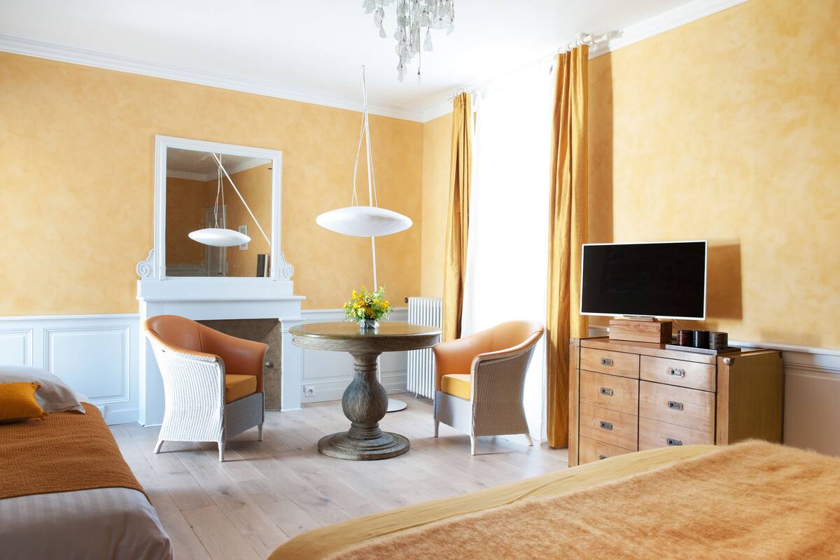 A hotel room with a bed, two chairs, a table with flowers, a TV on a wooden cabinet, and a large mirror.