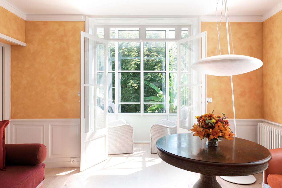 A room with a large open window, a round table with a vase of flowers, and two white chairs.