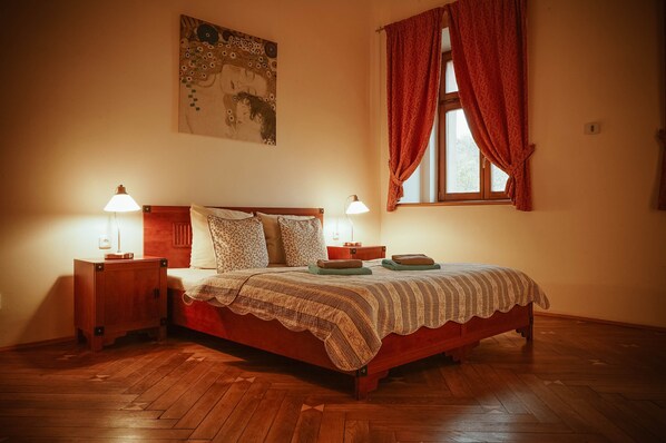 A bedroom with a wooden bed, two bedside tables with lamps, a window with red curtains, and a painting on the wall. - Hotel Stein Elbogen (Loket)