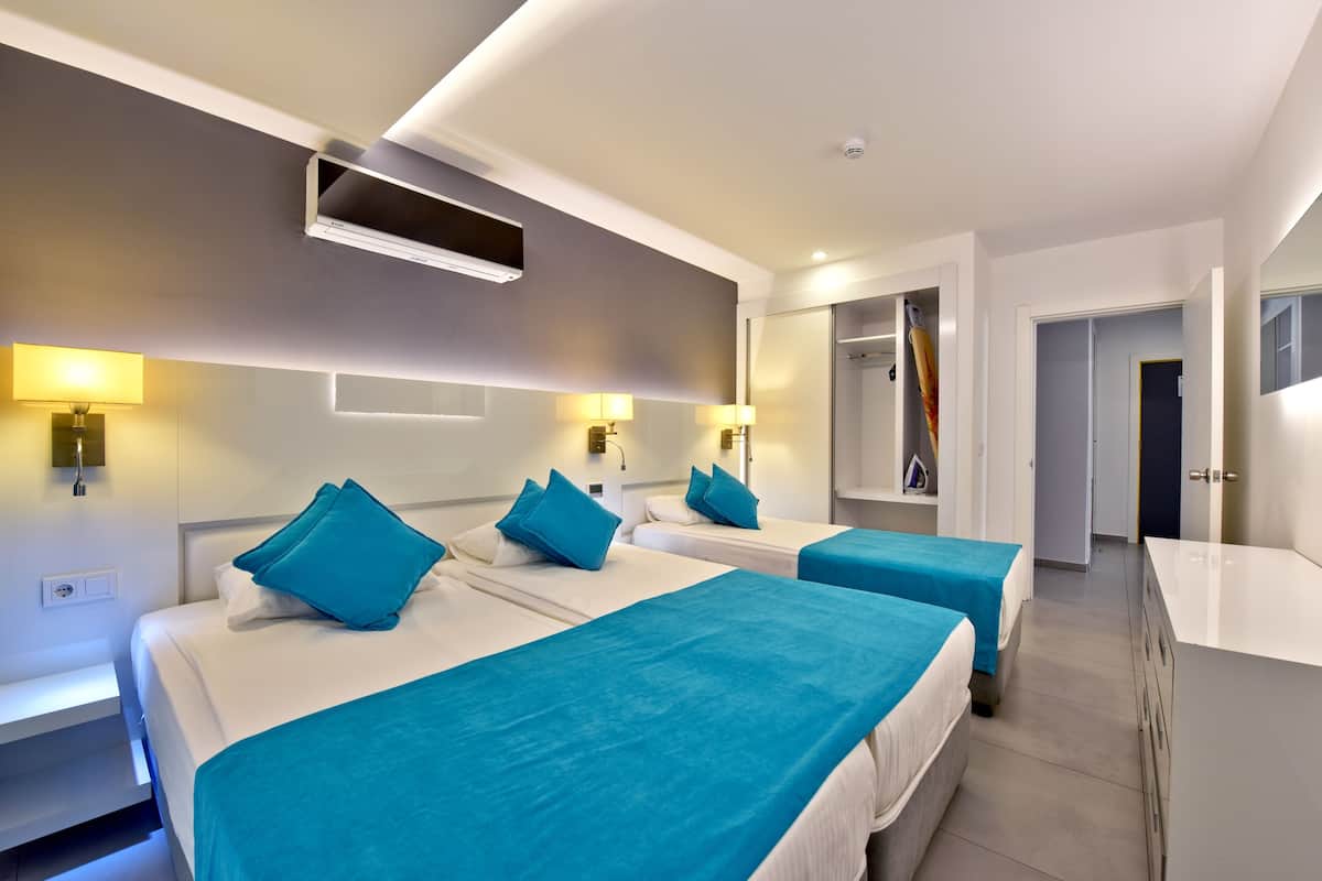 Una habitación de hotel con dos camas, cada una con cubrecama y almohadas azules, una mesita de noche blanca, aire acondicionado de pared y un espejo.