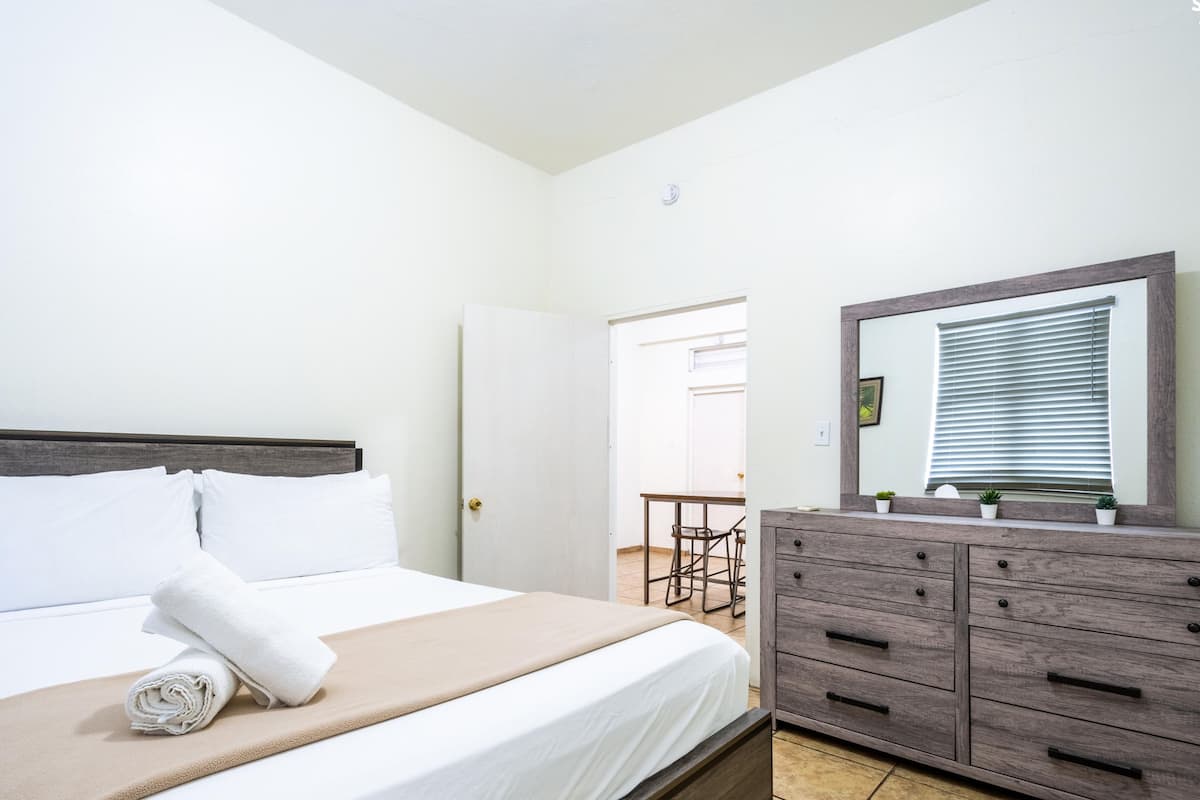 A hotel room with a bed, a dresser, a mirror, and a door leading to a dining area.