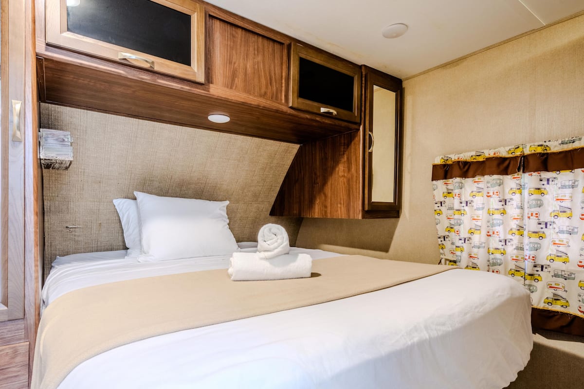 A neatly made bed with white linens and a rolled towel, wooden headboard, and a mirror above the bed.