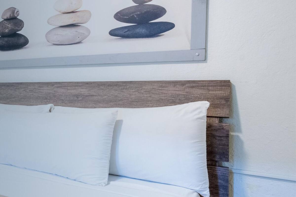A neatly made bed with white linens and a beige throw blanket, a wooden headboard, and a framed picture of stones on the wall above.