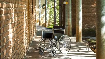 A wheelchair with a tray table and a cup holder, parked on a brick-paved walkway under a covered area with columns.