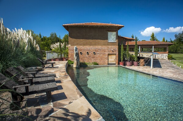 A swimming pool with a clear blue water surface, surrounded by a stone deck and a wooden bench area.