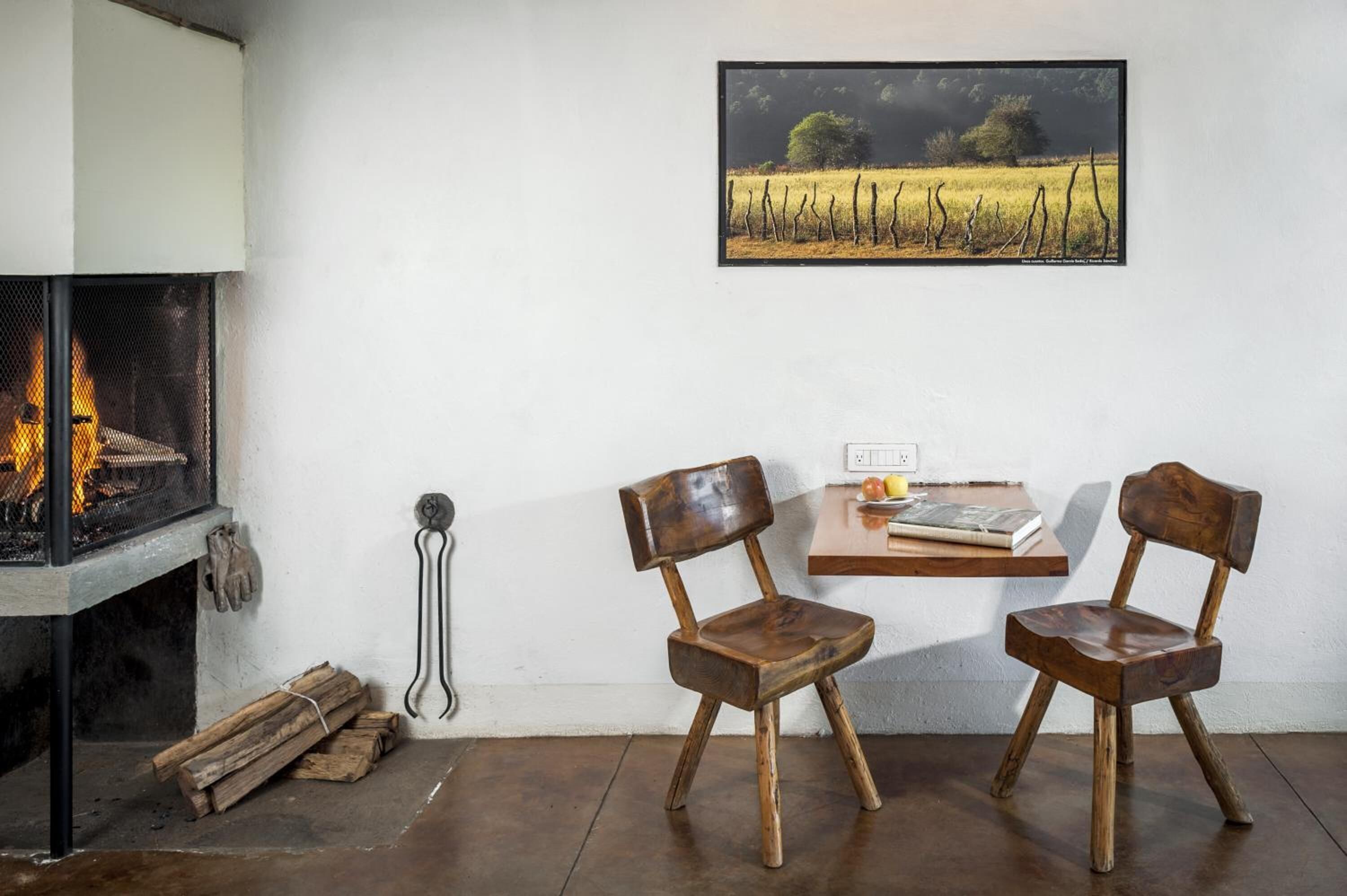 Una habitación acogedora con chimenea, dos sillas de madera, una mesa con libros y una vela, y un cuadro de paisaje enmarcado en la pared.