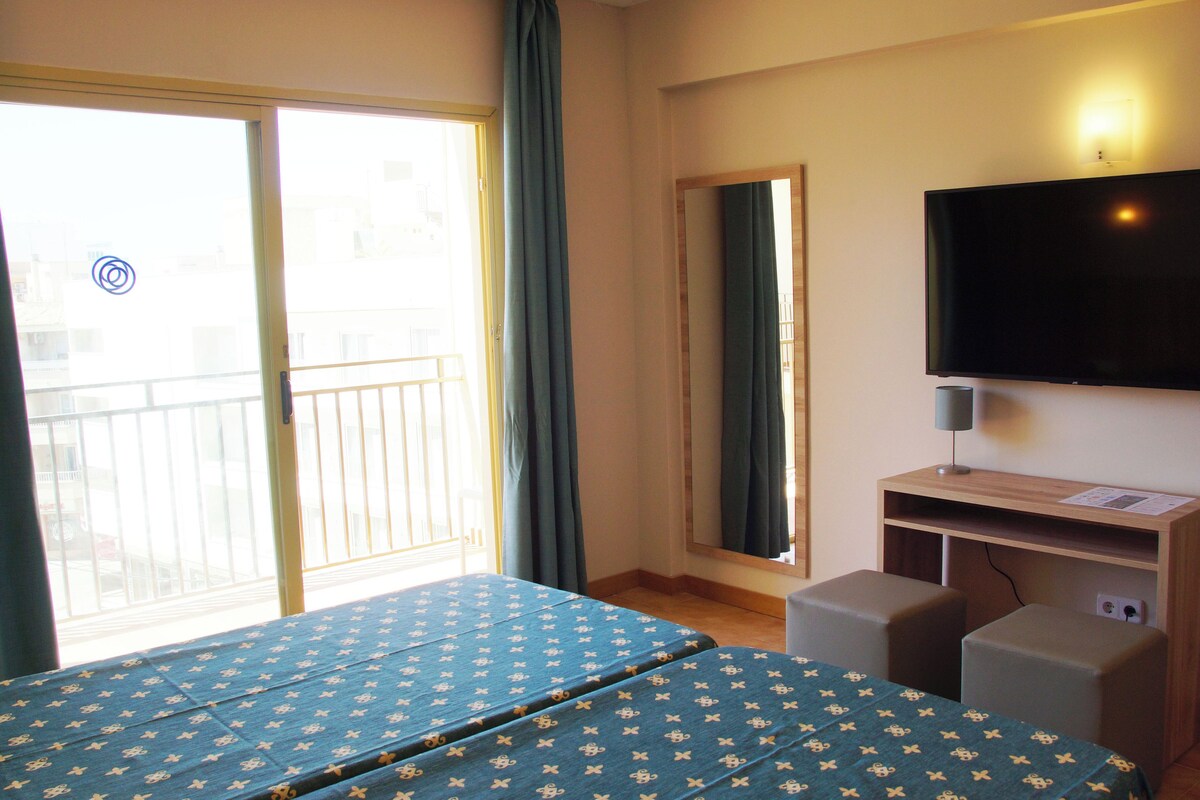 Quarto de hotel com cama, televisão, escrivaninha e varanda com cortinas.