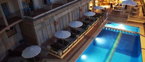 A hotel pool area with multiple lounge chairs and umbrellas, a second-floor balcony with railings, and a well-lit interior.