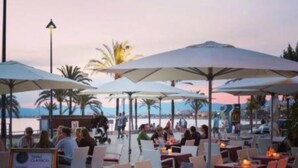Een buitenterras met tafels en stoelen onder parasols, met uitzicht op een strand.