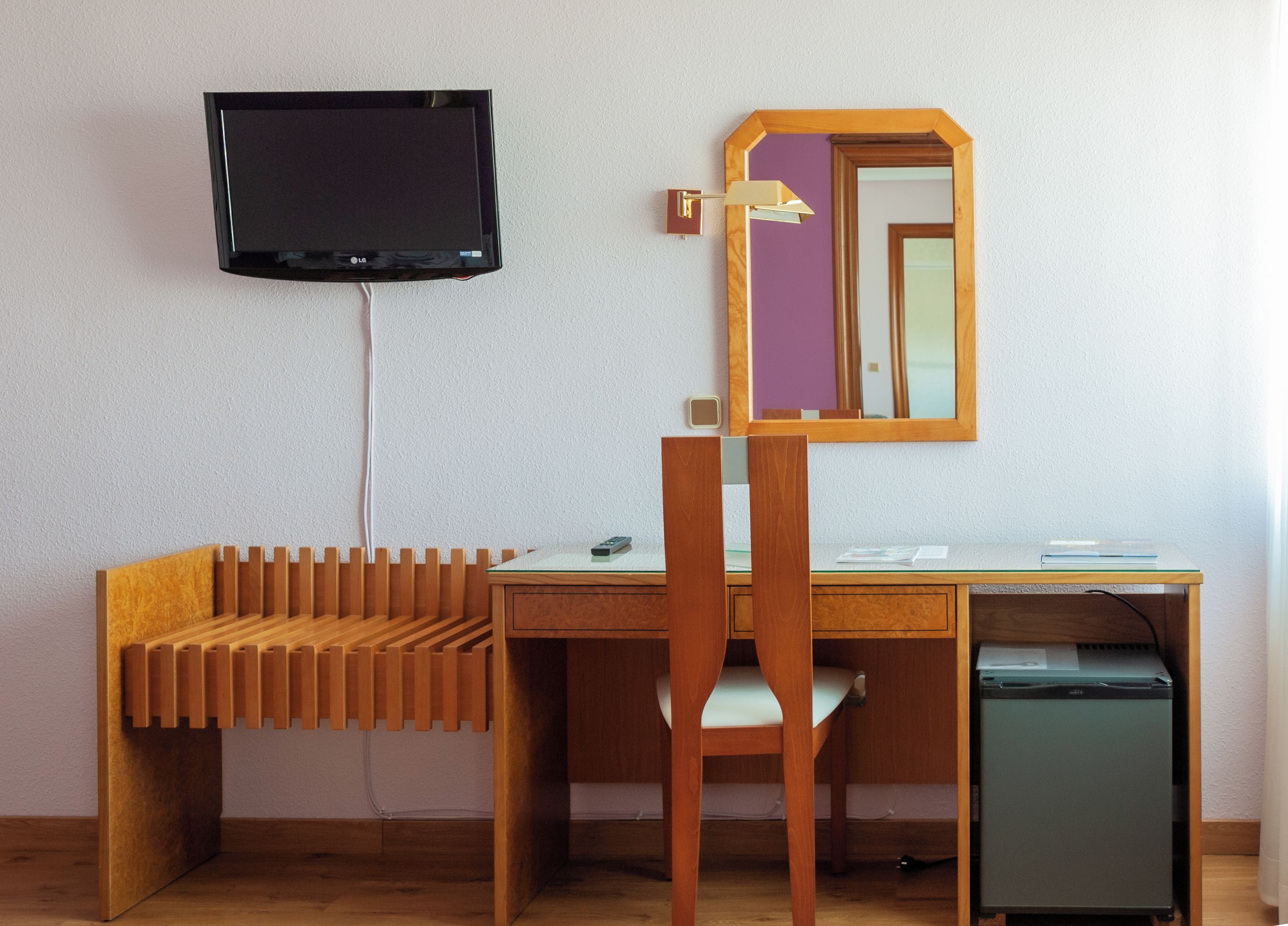 Una habitación con un escritorio de madera, una silla, un televisor montado en la pared, un espejo y un refrigerador pequeño.