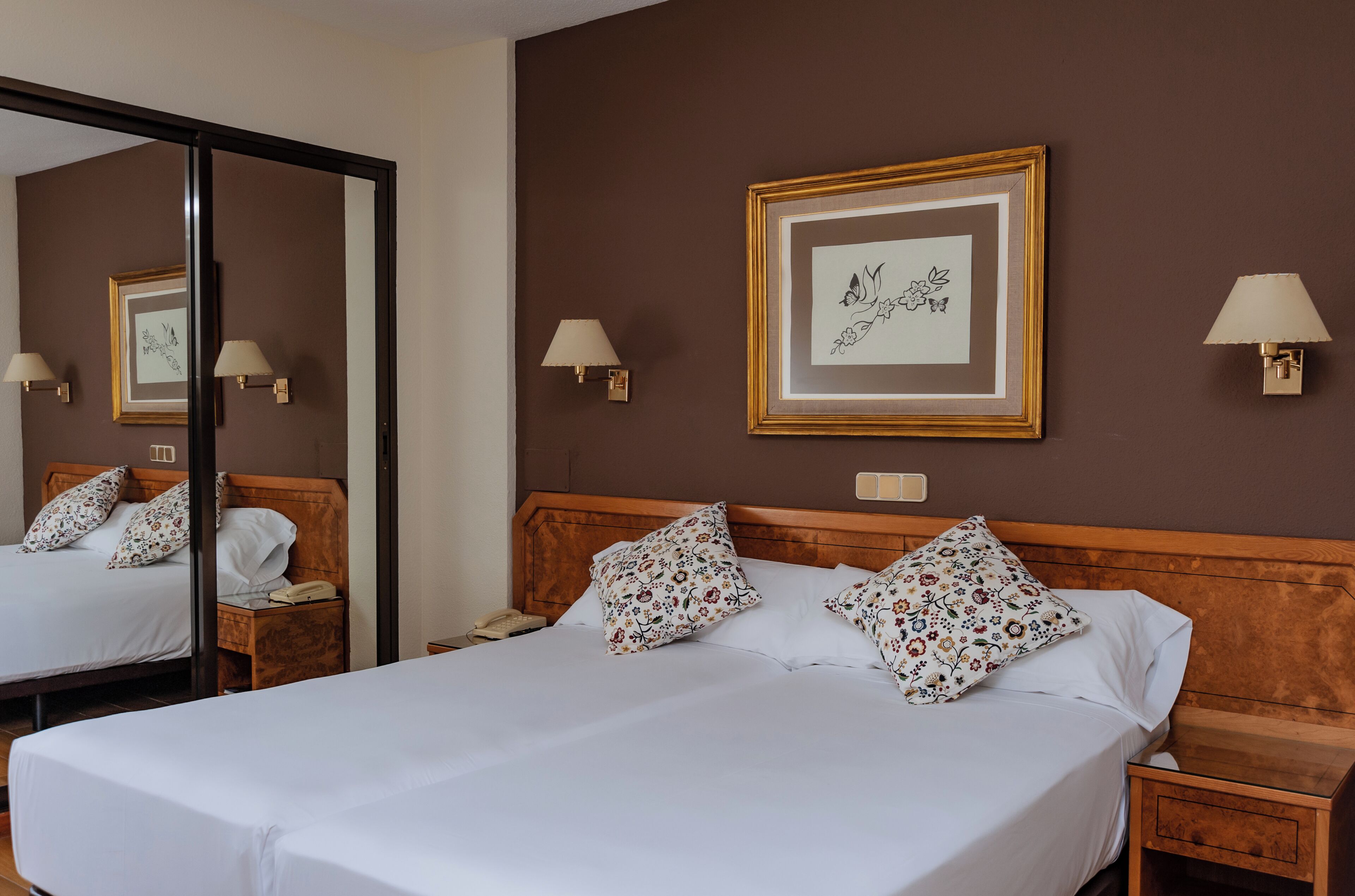Ein Hotelzimmer mit einem großen Bett, einem hölzernen Kopfteil, zwei Nachttischen mit Lampen, einem Spiegel und einem gerahmten Bild an der Wand.