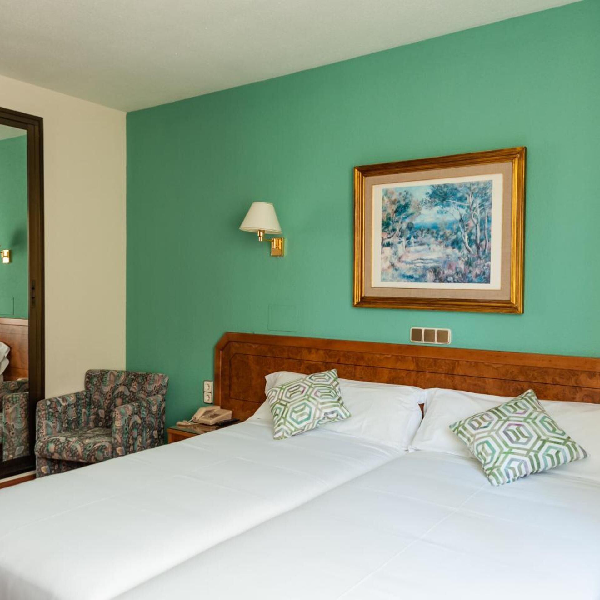 Habitación de hotel con cama, dos almohadas estampadas, mesita de noche con lámpara, un cuadro enmarco y una silla.