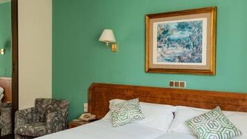 Habitación de hotel con cama, dos almohadas estampadas, mesita de noche con lámpara, un cuadro enmarco y una silla.