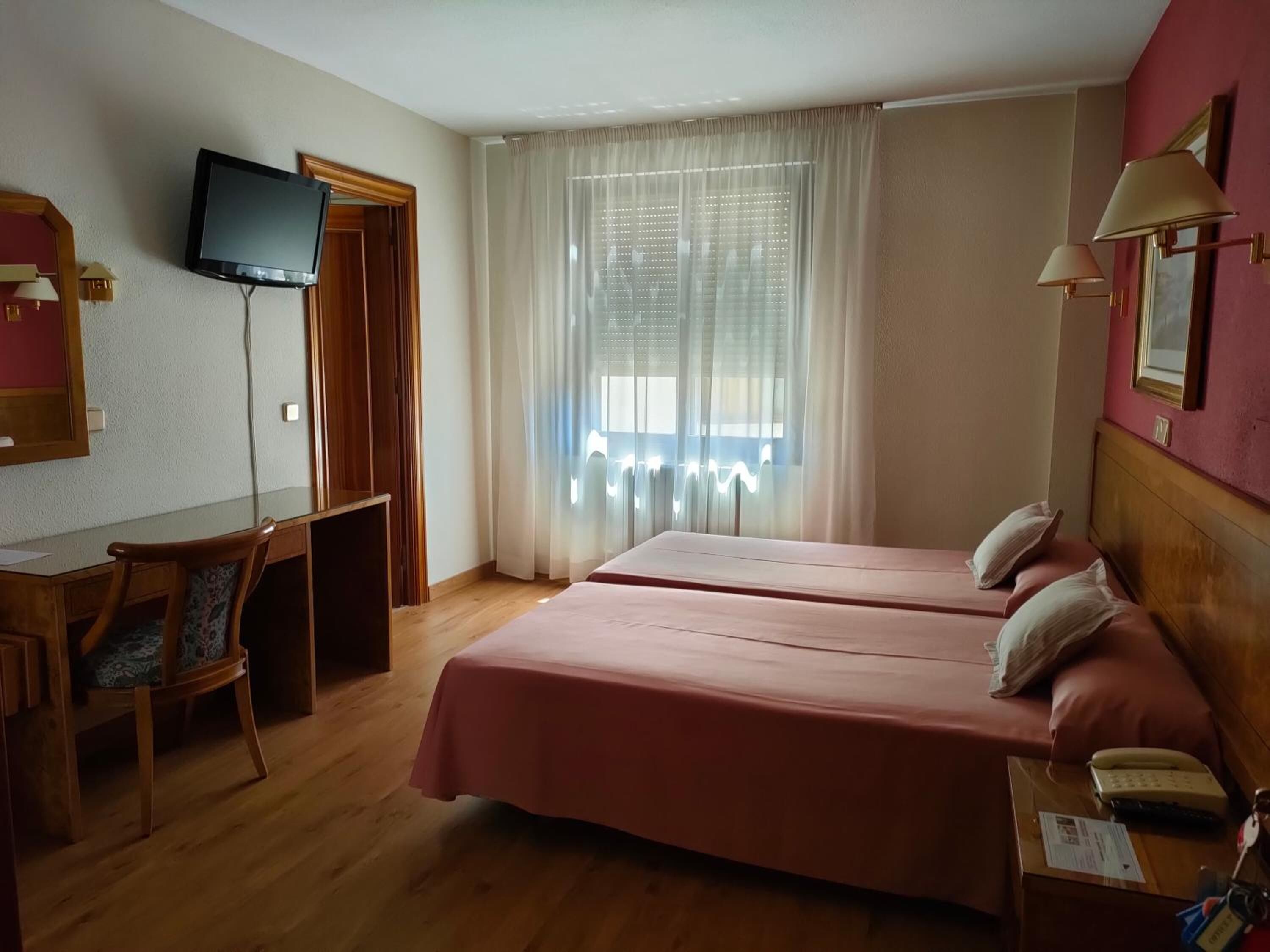 Habitación de hotel con dos camas, un escritorio, una silla, televisión y ventana con cortinas.