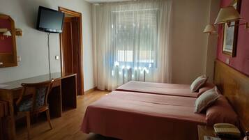 Habitación de hotel con dos camas, un escritorio, una silla, televisión y ventana con cortinas.