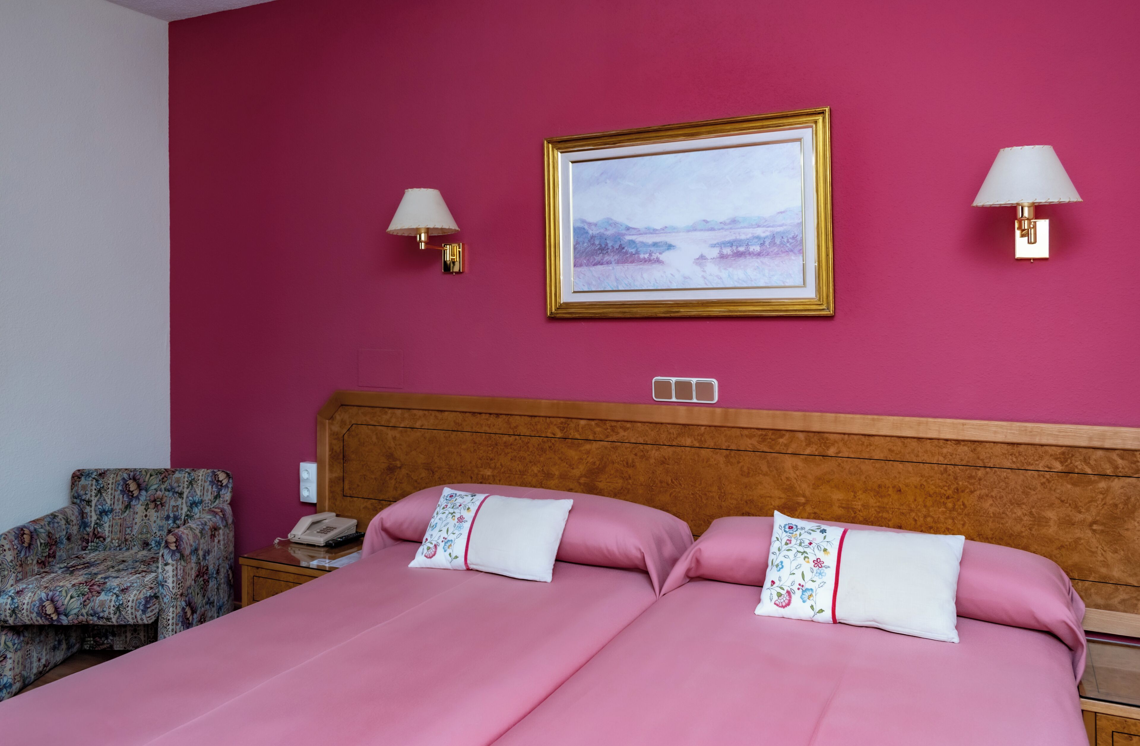 Habitación de hotel con cabecera de madera, dos camas con ropa de cama rosa, un sillón estampado con flores, un cuadro en la pared y dos lámparas de pared.