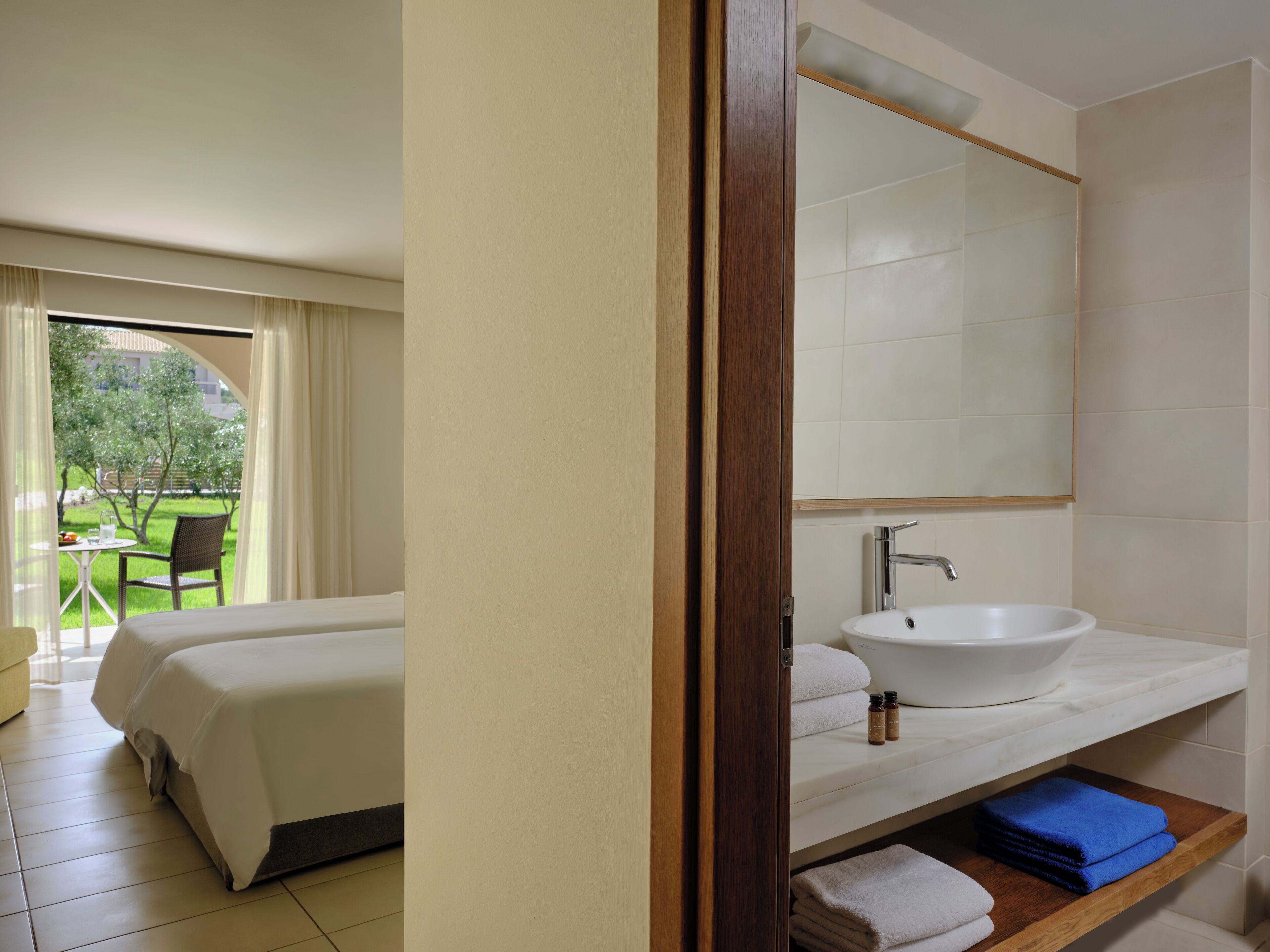 Une chambre d’hôtel avec un lit, une salle de bain dotée d’un grand miroir et d’un lavabo, et une vue sur un jardin depuis la fenêtre.