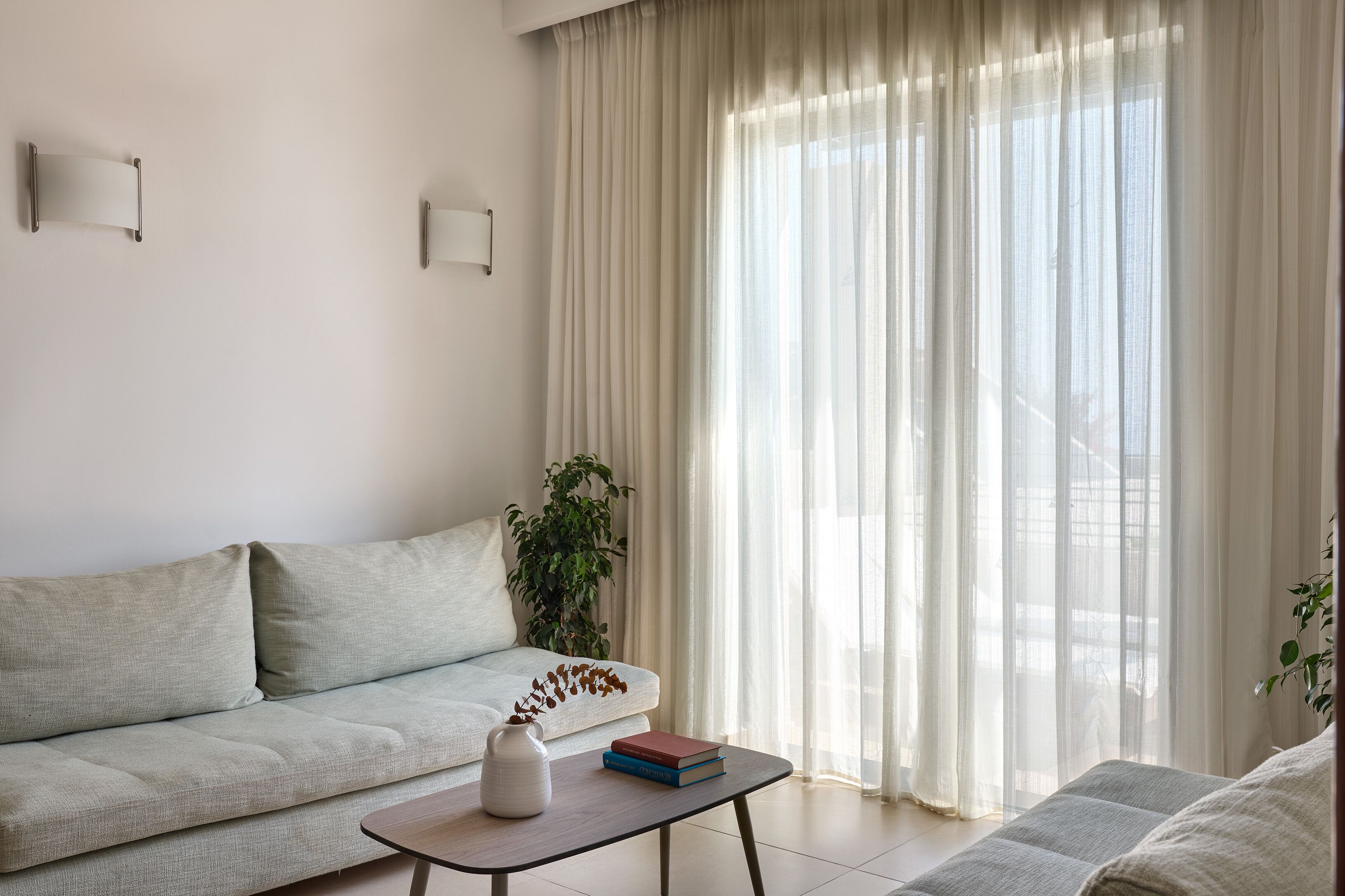 Una sala de estar con un sofá de color claro, una mesita con un florero y libros, y cortinas ligeras.