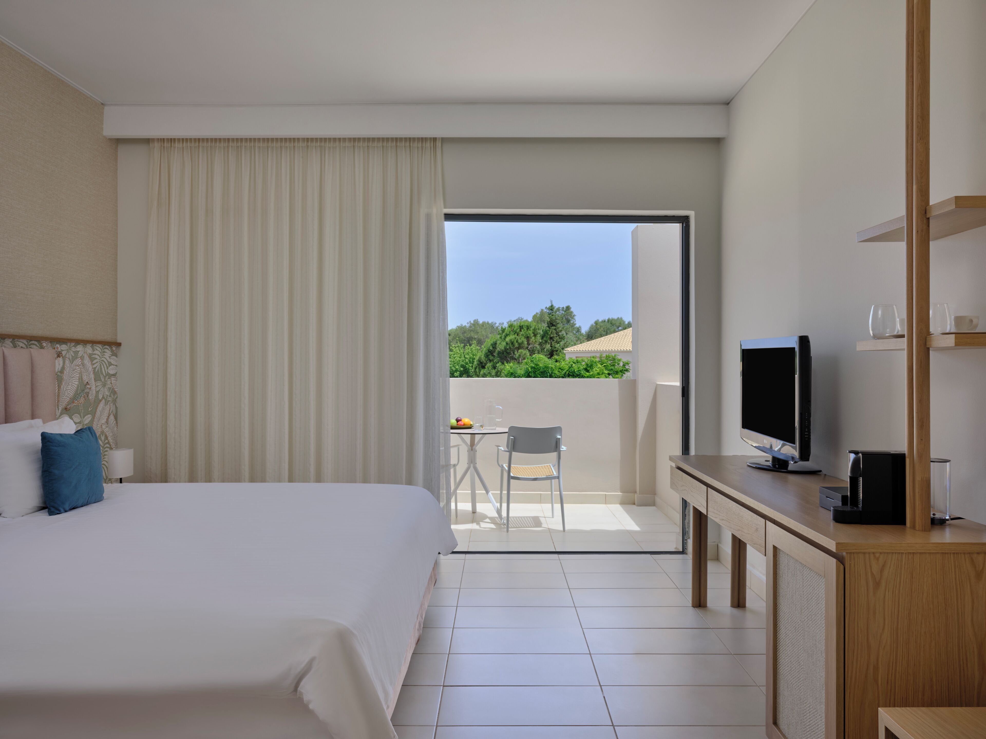 A hotel room with a bed, a desk with a TV, a balcony with a table and chair, and a view of greenery.
