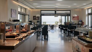A modern dining area with a buffet setup, seating arrangements, and large windows offering a view. - Atlantica Eleon Grand Resort - All Inclusive (Zakynthos)