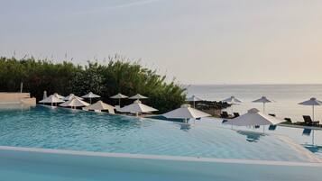 En pool med vita parasoller och solstolar, omgiven av grönska och med utsikt över havet.