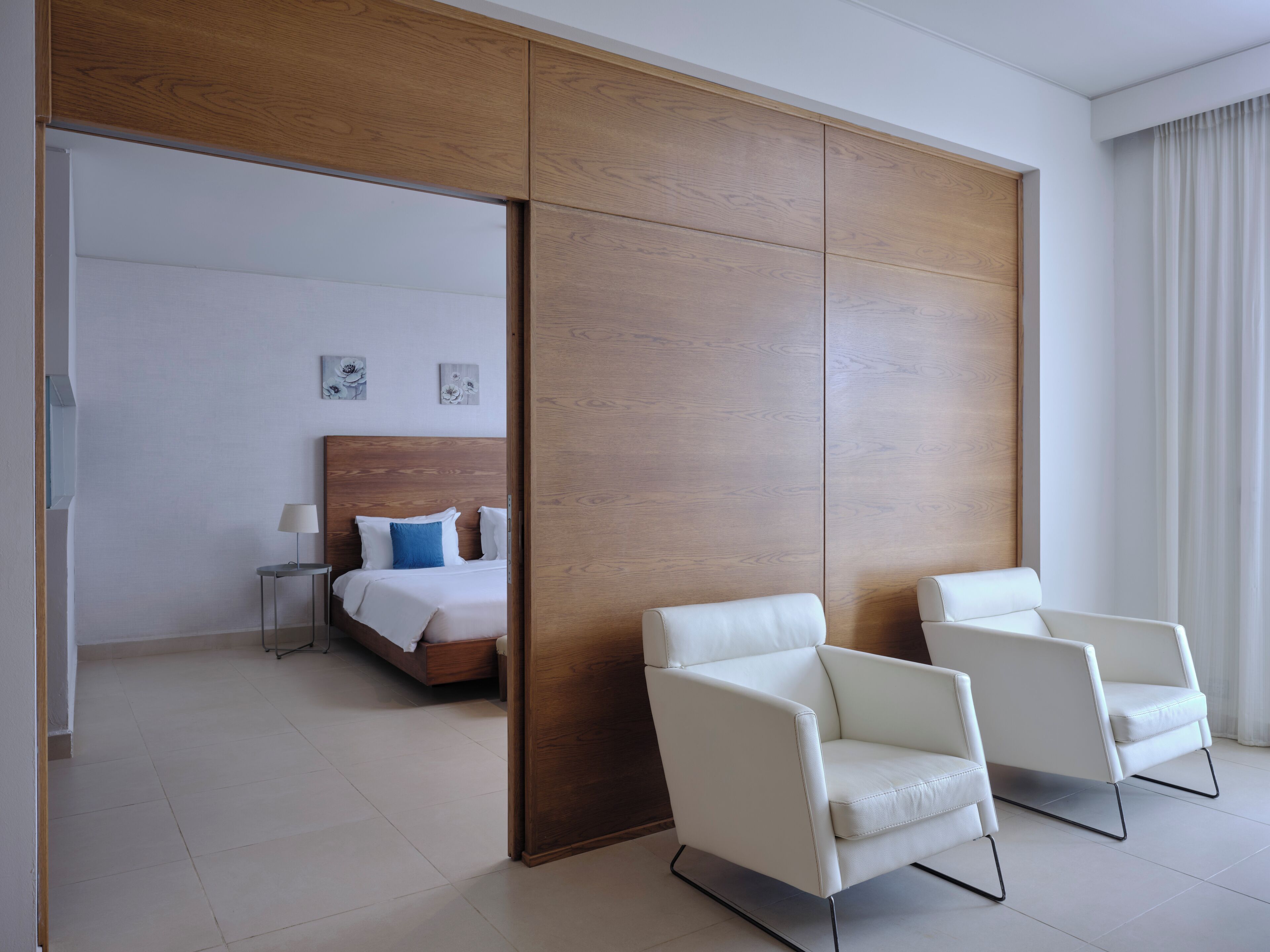 A hotel room with a wooden wardrobe, a bed with white and blue bedding, and two white armchairs.