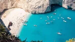 A coastal scene with boats on turquoise water, a sandy beach, and rocky cliffs. - Atlantica Eleon Grand Resort - All Inclusive (Zakynthos)