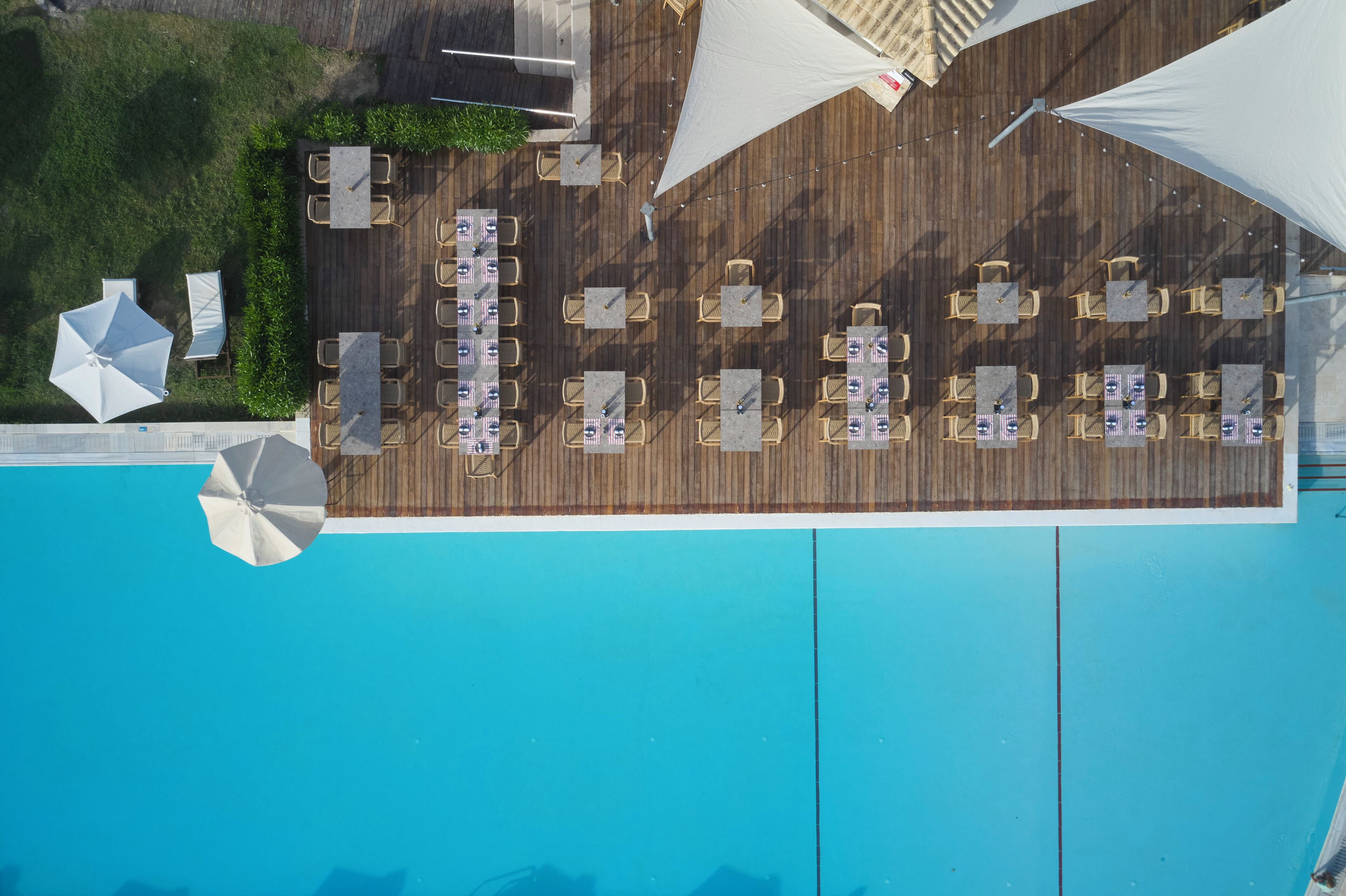 An aerial view of a poolside area with multiple tables and chairs, umbrellas, and a wooden deck.