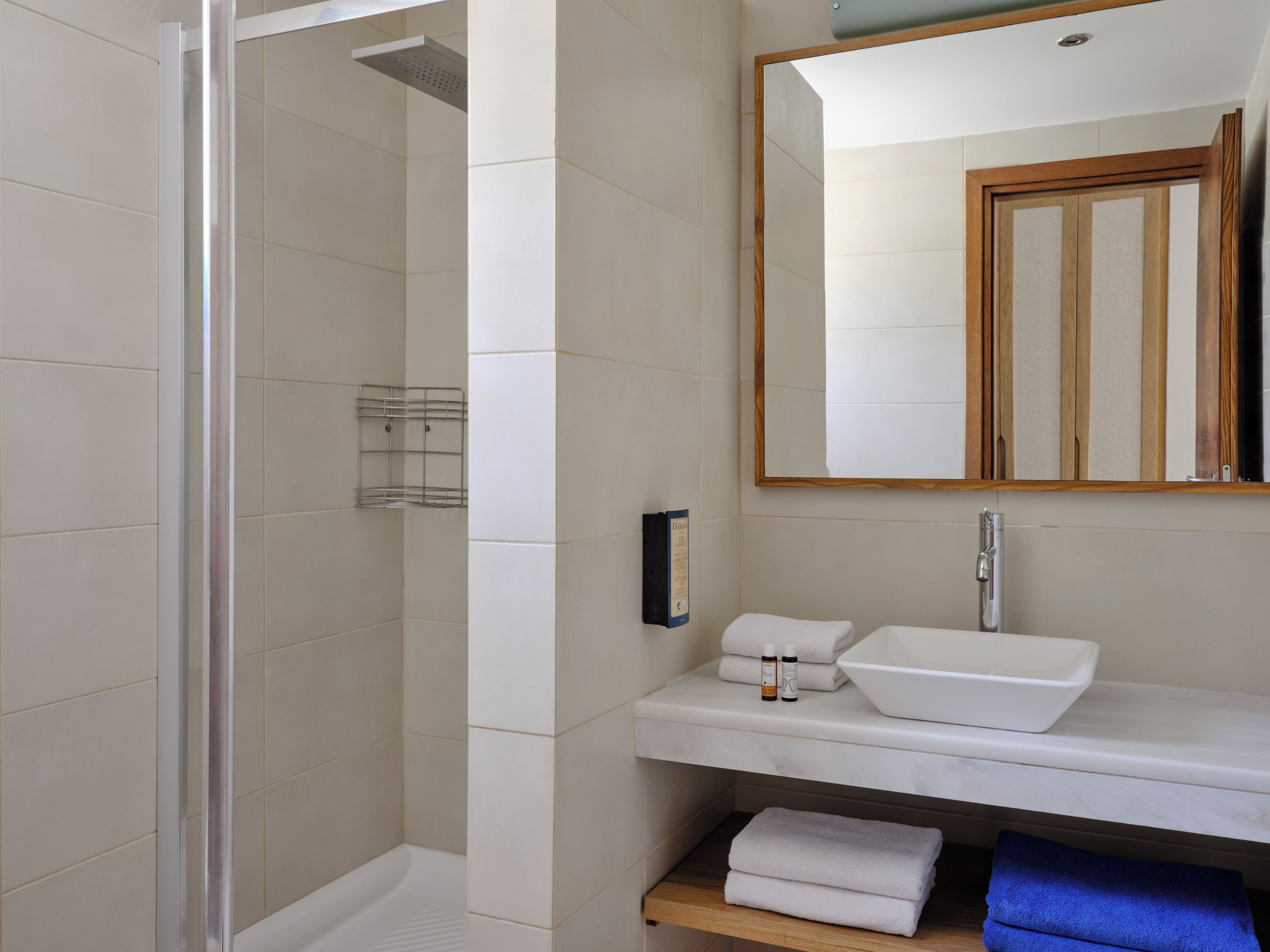 Un baño con cabina de ducha de vidrio, un lavamanos blanco con un espejo encima y una repisa con toallas y artículos de aseo.