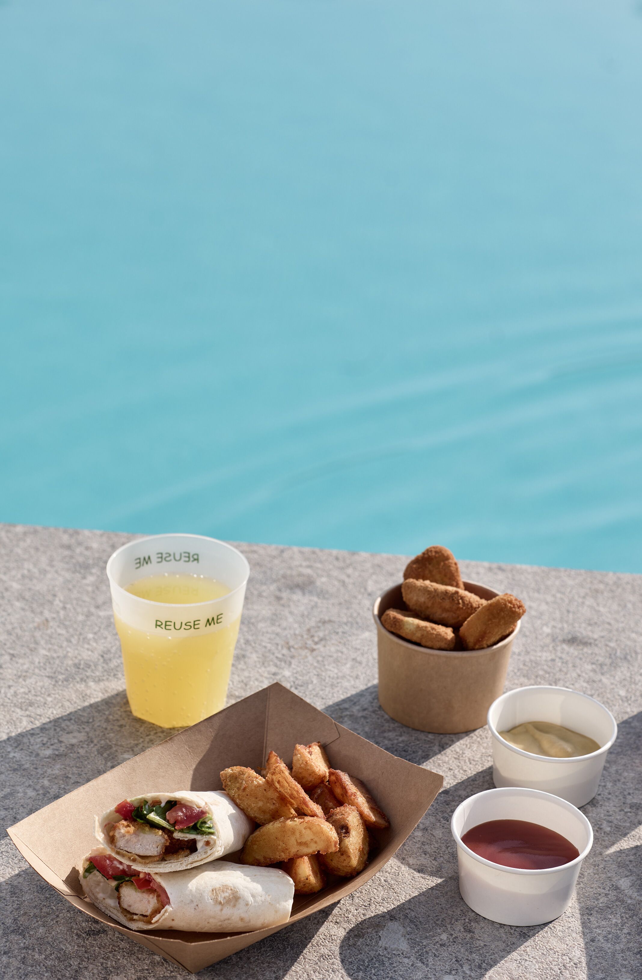 A meal consisting of a sandwich, fried items, and two drinks on a table by a pool.